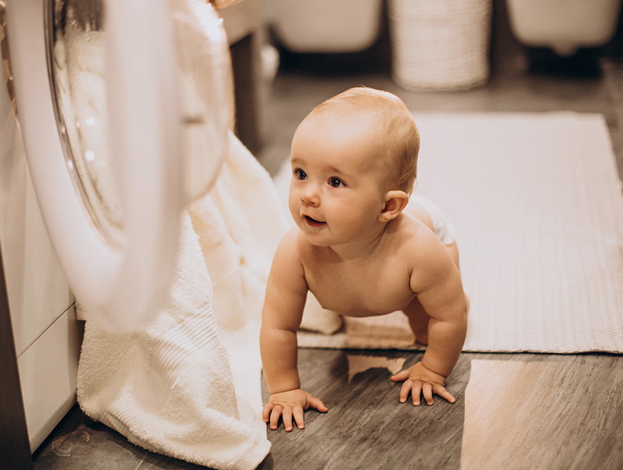 Este bebé está gateando junto a la lavadora abierta del lavadero de casa. Hay una toalla blanca en su interior. El bebé lleva un pañal y nada más, debe ser verano.
