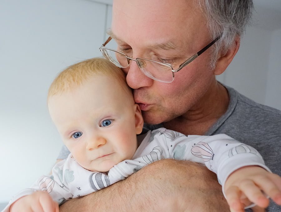 Este abuelo le está dando un beso en la oreja a su nieto pequeño que tiene cogido en brazos. Es un bebé varón.