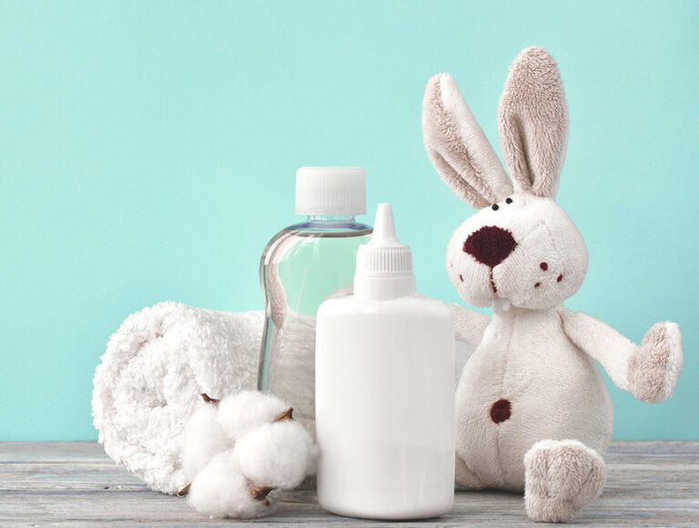 Varios productos de higiene y cuidados del bebé sobre una mesa de madera. Hay aceite corporal, loción, una toalla y un peluche de conejo.