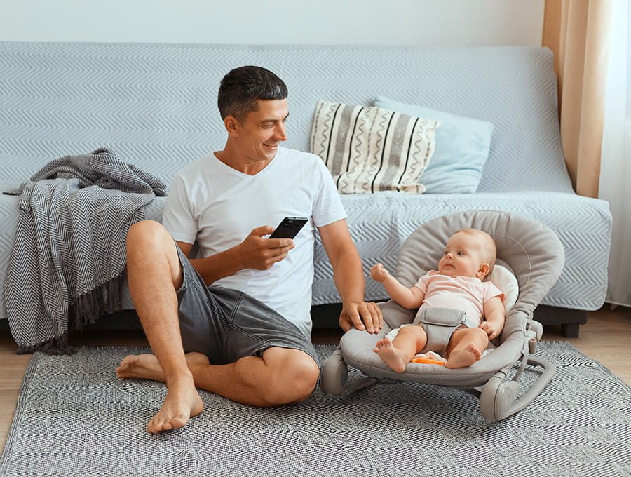 Este padre está sentado en la alfombra del salón junto a su bebé. El pequeño está tumbado en una mecedora y se lo pasa genial. El padre le va a sacar una foto con el teléfono.