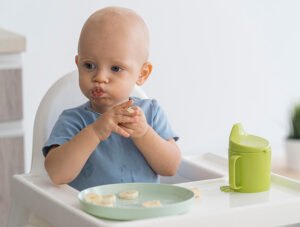 Este niño pequeño está sentado en su trona junto a la mesa. Está comiendo unas rodajas de plátano que le ha cortado su padre.