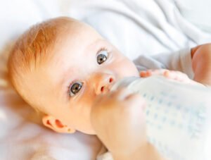 Este bebé está tomando un poco de leche en el biberón. Está tumbado boca arriba en la cuna.