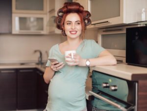 Esta mujer embarazada tiene rizadores (rulos) de pelo en la cabeza. Parece que se va a hacer la permanente en casa. Ahora está tomando un café en la cocina mientras mira el móvil.