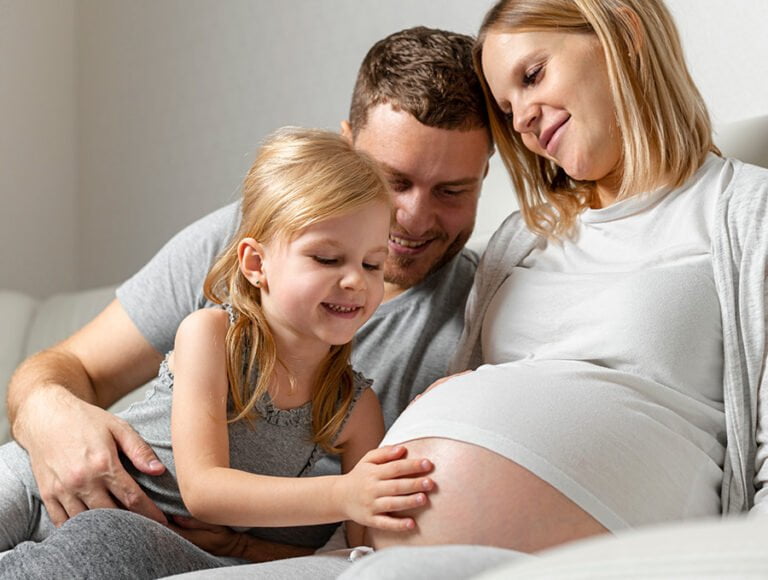 Embarazada está sentada en el sofá. Su hija está tocando su barriga, mientras que el marido las observa y se rie.
