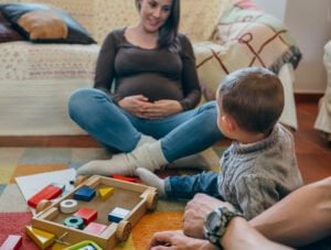 Esta embarazada está sentada en la alfombra junto al sofá. Su hijo la está mirando y se rien.