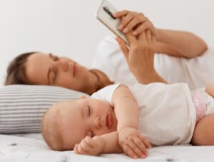Esta madre está tendida en la cama junto a su bebé. Ella mira el móvil y el crío duerme plácidamente.