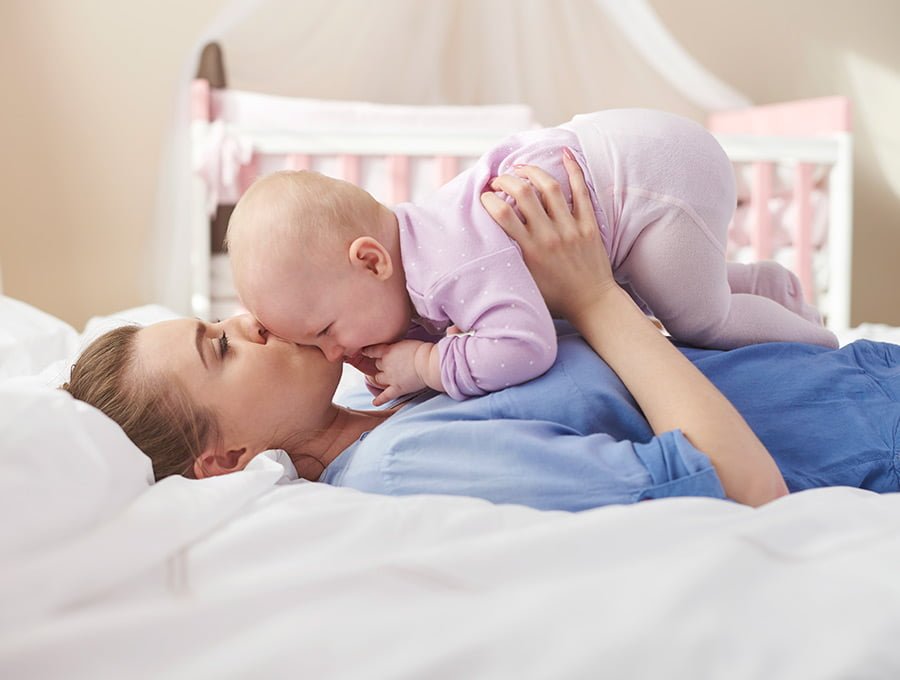 Esta madre tiene a su bebé sombre el pecho. Están tumbados en la cama y la madre le da un beso en la cara. Hay una cuna al fondo.