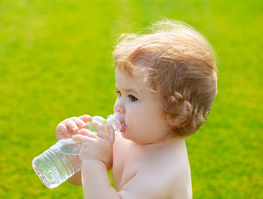 Este niño pequeño está bebiendo agua de una botella de agua de plástico. Está sobre el césped del jardín.