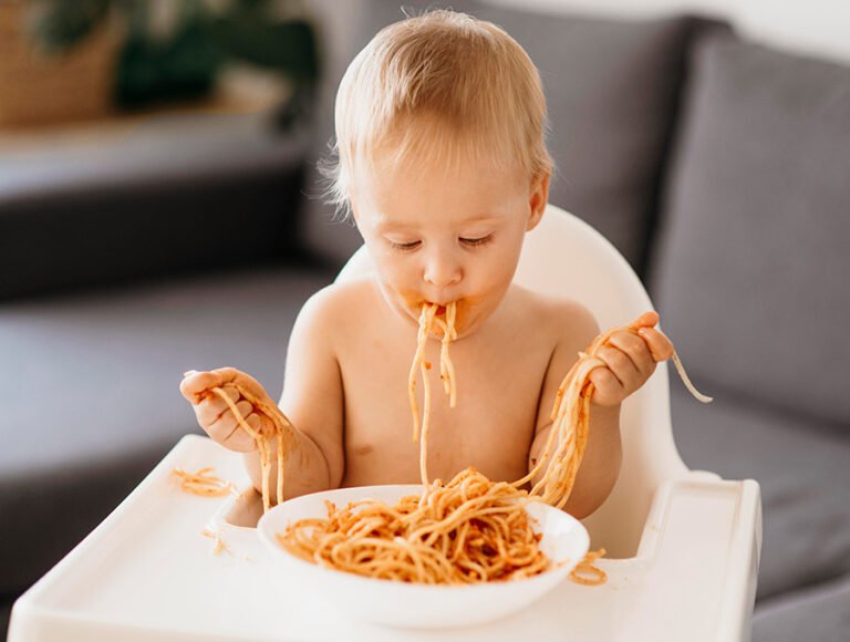 Este niño pequeño está sentado en la trona delante de la mesa. Está comiendo espaguettis y lo está manchando todo.