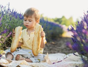Este niño está comiendo una barra de pan. Está sentado sobre una manta.