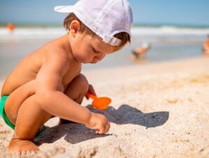 Este niño pequeño está jugando con la arena en la orilla del mar. Lleva una gorra blanca y un bañador verde.