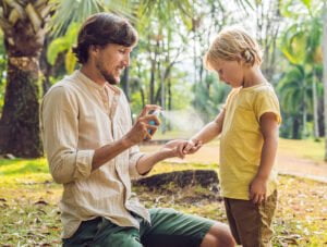 Este padre le está echando un poco de spray antimosquitos con citronela a su hijo pequeño. Están en una selva con palmeras y otras plantas tropicales.