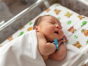 Este recién nacido está durmiendo plácidamente en una cuna del hospital. Está tapado con una mantita blanca.