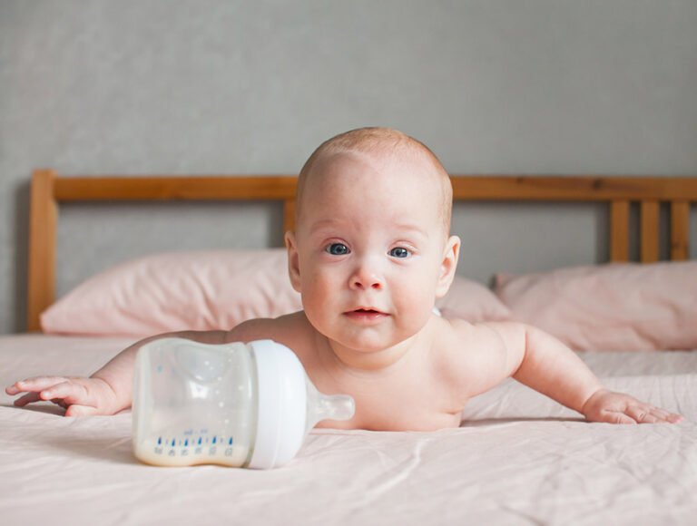 Este bebé está gateando encima de la cama. Tiene el biberón de leche también a su lado.