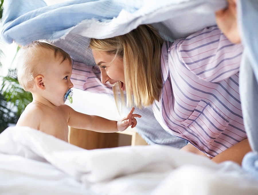 Esta madre está jugando con el edredón y su bebé encima de la cama. El pequeño lleva puesto un chupete nuk.