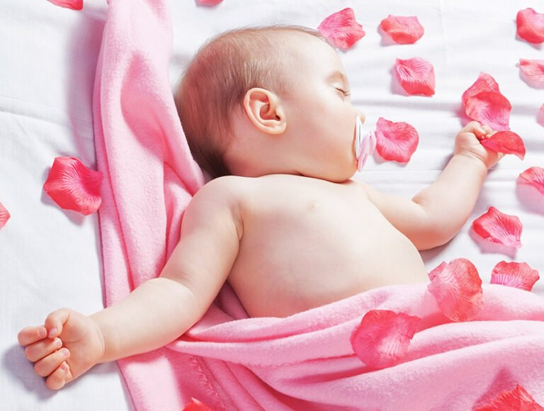 Este bebé está durmiendo. Está tapado con una manta rosa y varios pétalos de rosa decorativos.