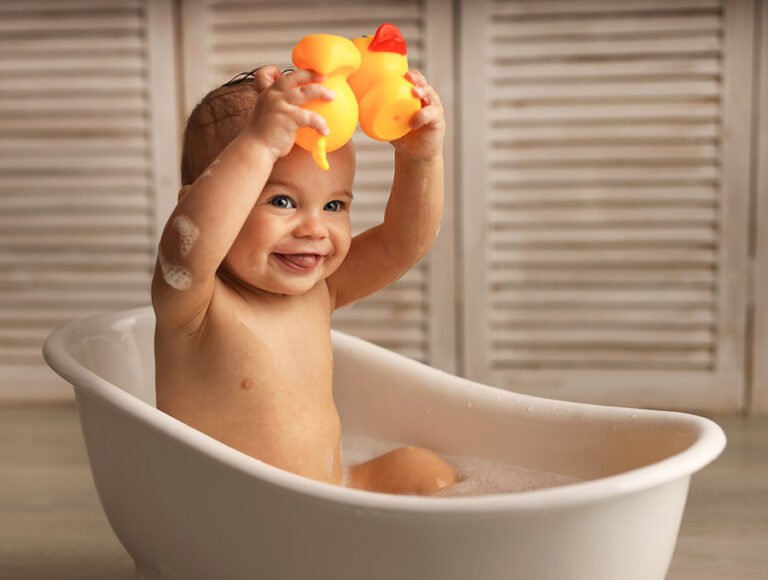 Este bebé está dentro de una bañera pequeña. Se está dando un baño con sus patitos de goma amarillos.