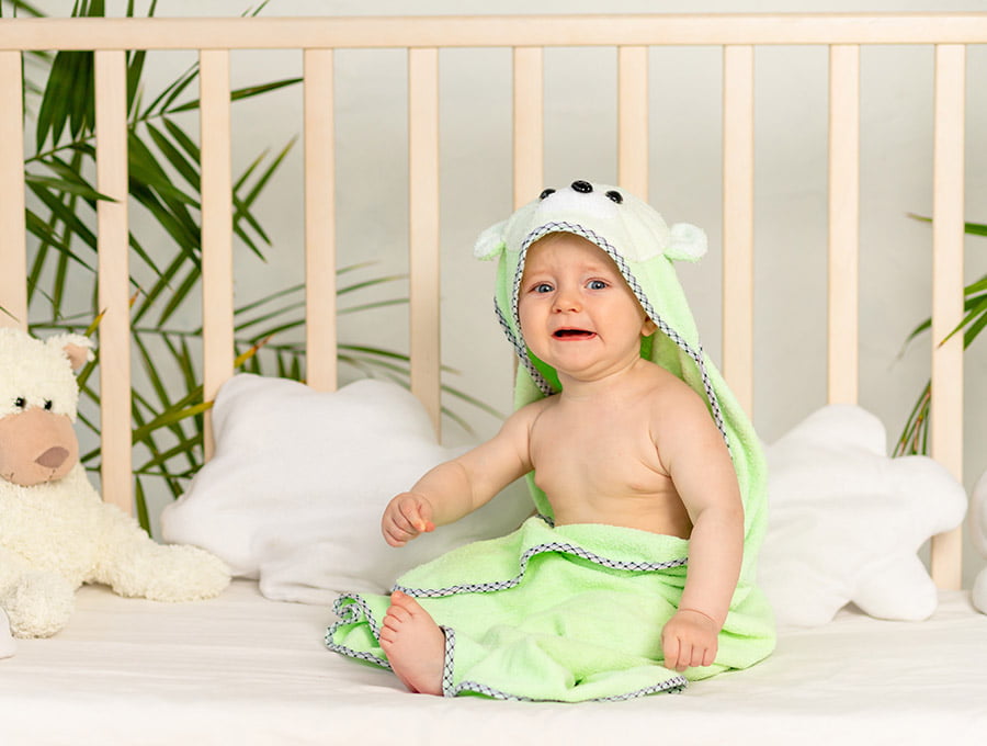Este bebé se ha dado un baño. Ahora está en su cuna secándose con la toalla de animales. Está medio llorando.