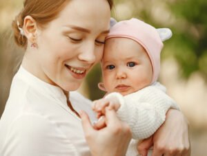 Bebé niña con un sombrero rosa. La madre la tiene cogida en brazos. Le toca la mano pequeñita.