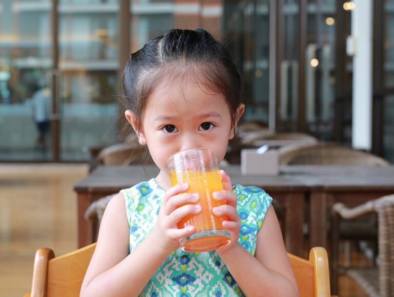 Esta niña pequeña se está tomando un zumo de naranja en un vaso de cristal.