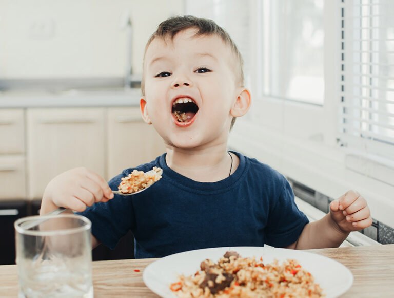 Este niño pequeño está comiendo un plato de arroz con la cuchara. Está sentado en la mesa de la cocina. Tiene la boca abierta para enseñar que está comiendo.