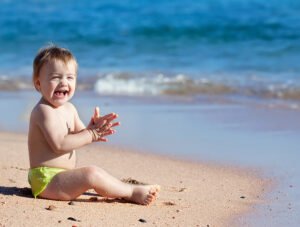 Este niño pequeño está sentado en la arena de la playa junto a la orilla. Parece muy feliz.