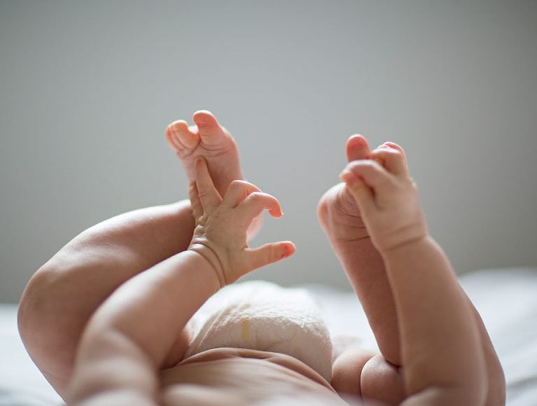 Bebé con pañal se toca los pies con las manos encima de la cama.