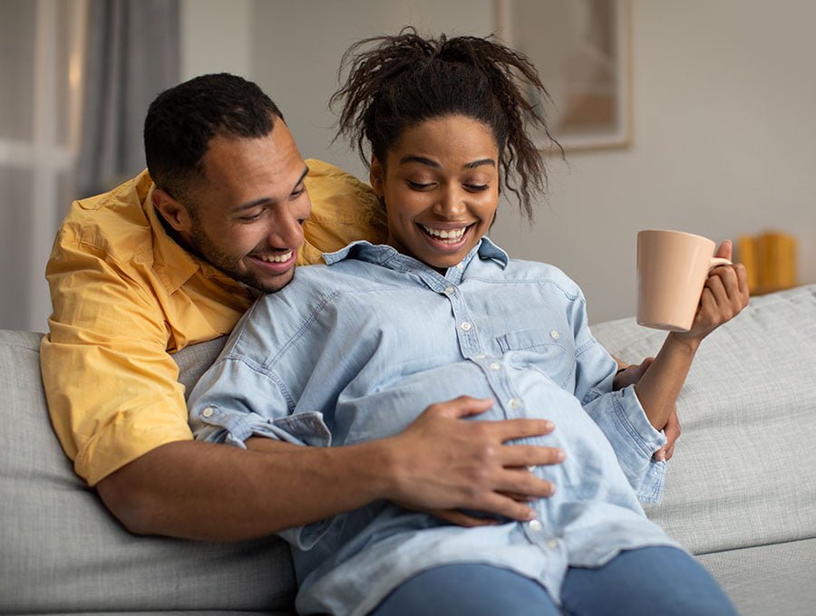 Embarazada se toma una taza de té mientras el marido le toda la barriga y se ríen.